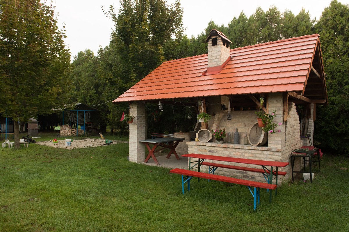 Kültéri, cserepes tetős sütő-főző ház sörpadokkal a kertben.